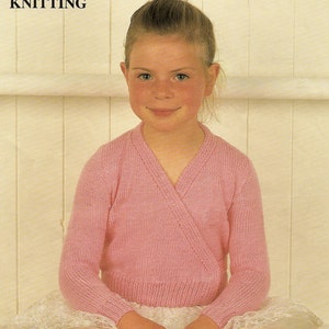 May include: A young girl wearing a pink knitted wrap cardigan with a V-neck and long sleeves. The cardigan is made with a double knit stitch. The girl is sitting on a white tulle skirt.