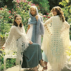 May include: Three women wearing crocheted shawls in a garden setting. The shawls are white, light blue, and cream with fringe detailing. The women are wearing dresses and skirts.
