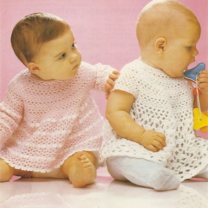 May include: Two babies wearing crocheted dresses. The baby on the left is wearing a pink dress and the baby on the right is wearing a white dress. Both babies are sitting on a pink surface.