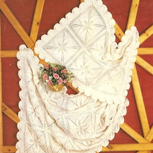 May include: Two off-white, crocheted blankets with a floral design and scalloped edges are displayed. A small basket of pink flowers sits between the blankets. The blankets are draped over a wooden structure with a red background.