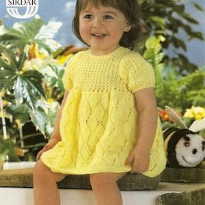 May include: A yellow knitted baby dress with a diamond pattern. The dress is worn by a young girl sitting on a stone wall. The girl is smiling and has short brown hair. The image includes the text "Snuggly DK" and "Sirdar" in the top left corner.