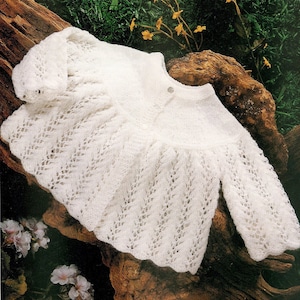May include: A white, knitted baby cardigan with a delicate, openwork pattern. The long-sleeved sweater features a scalloped hem and is buttoned at the neckline. The garment is displayed on a piece of weathered wood.