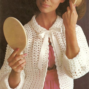 Peut inclure: Un cardigan en crochet blanc avec un nœud papillon en satin blanc. Le cardigan a un motif en dentelle et est ouvert sur le devant. Le mannequin porte une robe rose en dessous.