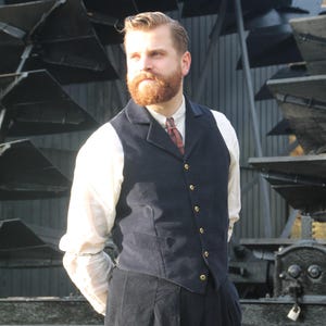 May include: A man in a navy blue vest with gold buttons, a white shirt, and dark pants. He has a red beard and is standing in front of a large, industrial-looking structure. The outfit has a vintage, classic style.