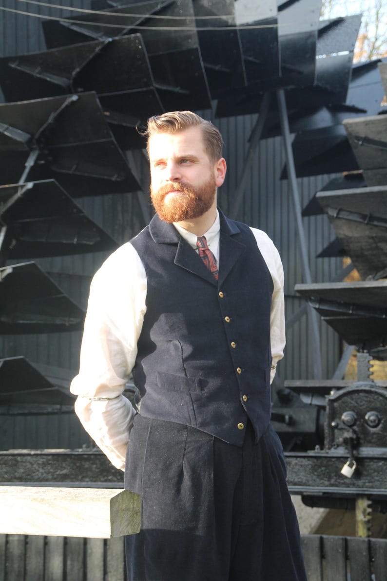 May include: A man in a navy blue vest with gold buttons, a white shirt, and dark pants. He has a red beard and is standing in front of a large, industrial-looking structure. The outfit has a vintage, classic style.