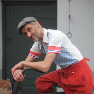 May include: A man wearing a white polo shirt with blue patterned panels and red and blue striped sleeves, paired with bright red pants. He is wearing a gray newsboy cap and leaning on a bicycle.
