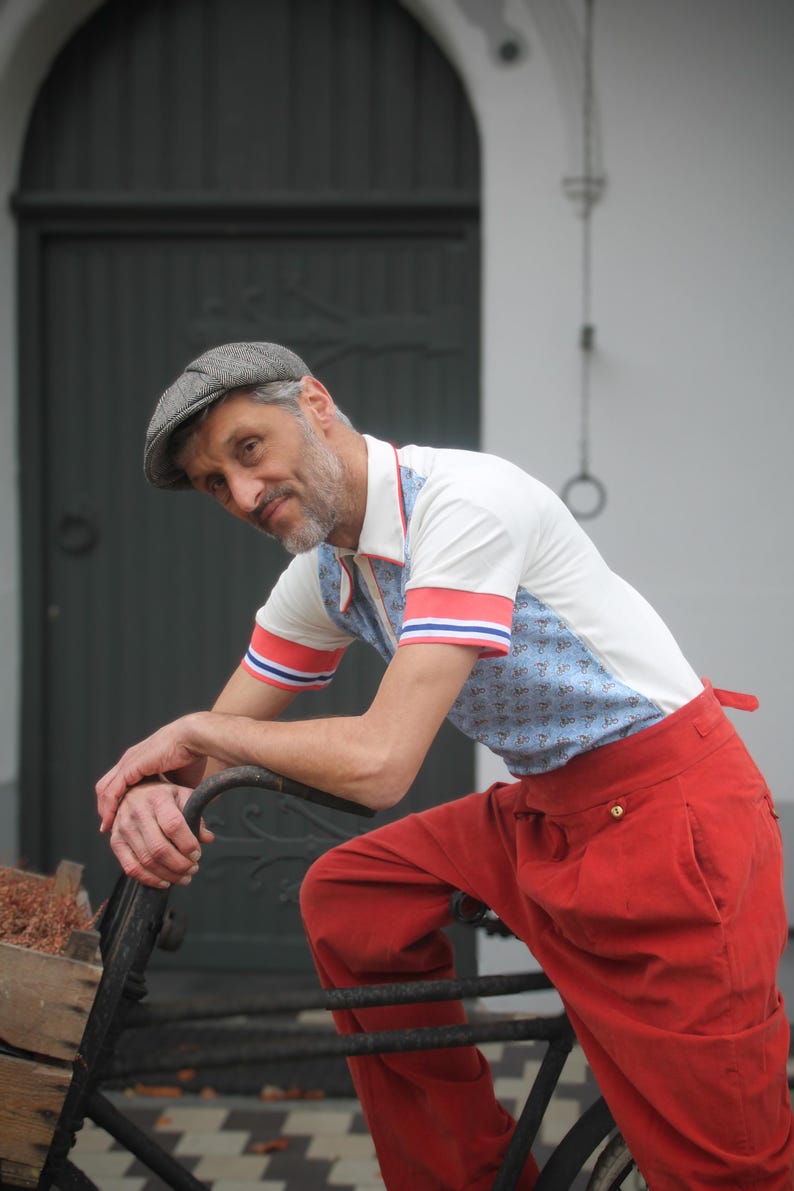 May include: A man wearing a white polo shirt with blue patterned panels and red and blue striped sleeves, paired with bright red pants. He is wearing a gray newsboy cap and leaning on a bicycle.