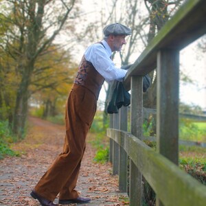 May include: A man in a brown corduroy suit, white shirt, and patterned vest leans on a wooden fence. He wears a gray cap and dark shoes. The scene is outdoors on a path covered in fallen leaves, with trees in the background.