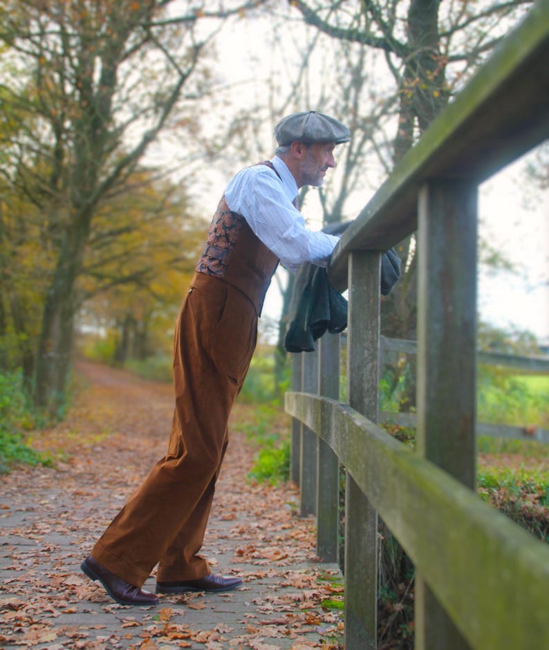 May include: A man in a brown corduroy suit, white shirt, and patterned vest leans on a wooden fence. He wears a gray cap and dark shoes. The scene is outdoors on a path covered in fallen leaves, with trees in the background.