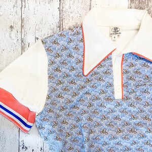 May include: A short-sleeved polo shirt with a blue pattern of bicycles on a light blue background. The shirt has white sleeves and a white collar with red trim. The sleeve cuffs have red and blue stripes.