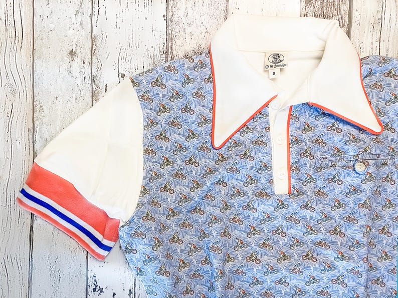 May include: A short-sleeved polo shirt with a blue pattern of bicycles on a light blue background. The shirt has white sleeves and a white collar with red trim. The sleeve cuffs have red and blue stripes.