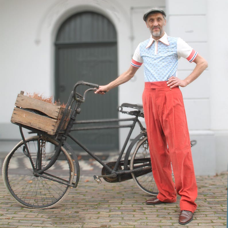 May include: An old black bicycle with a wooden crate. The bicycle is parked on a brick surface. The person is wearing a light blue and white patterned polo shirt, red trousers, and a grey cap.