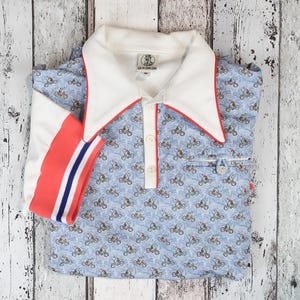 May include: A light blue polo shirt with a bicycle pattern. The shirt has a white collar with red trim, a white placket, and a red and blue striped sleeve cuff. The shirt is folded on a white wooden surface.
