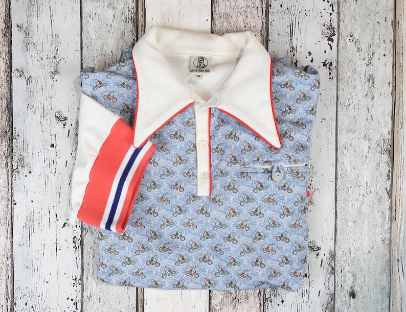 May include: A light blue polo shirt with a bicycle pattern. The shirt has a white collar with red trim, a white placket, and a red and blue striped sleeve cuff. The shirt is folded on a white wooden surface.