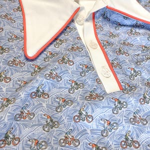 May include: A light blue polo shirt featuring a pattern of cyclists on bicycles. The shirt has a white collar with a coral-colored trim and a white button placket. The background has a wavy pattern.