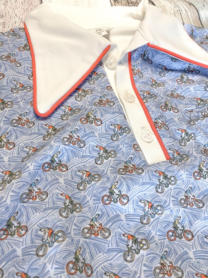 May include: A light blue polo shirt featuring a pattern of cyclists on bicycles. The shirt has a white collar with a coral-colored trim and a white button placket. The background has a wavy pattern.