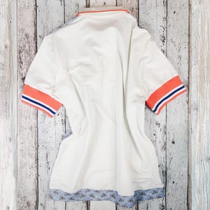 May include: White polo shirt with orange and blue striped sleeve cuffs and collar trim. The bottom hem features a blue floral pattern. The shirt is laid flat on a light-colored wooden surface.