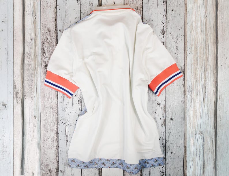 May include: White polo shirt with orange and blue striped sleeve cuffs and collar trim. The bottom hem features a blue floral pattern. The shirt is laid flat on a light-colored wooden surface.