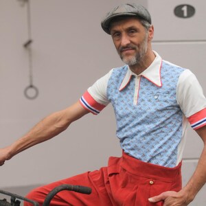 May include: A man wearing a vintage-style outfit, including a light blue polo shirt with a small pattern, red and white accents, and red trousers. He is wearing a gray cap and is holding a bicycle handlebar.