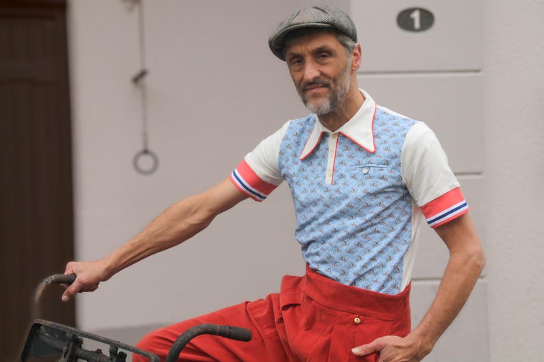 May include: A man wearing a vintage-style outfit, including a light blue polo shirt with a small pattern, red and white accents, and red trousers. He is wearing a gray cap and is holding a bicycle handlebar.