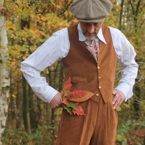 A brown suede waistcoat with gold buttons, worn with matching trousers. The outfit includes a white striped shirt, a patterned tie, and a tweed cap. The person is holding autumn leaves.