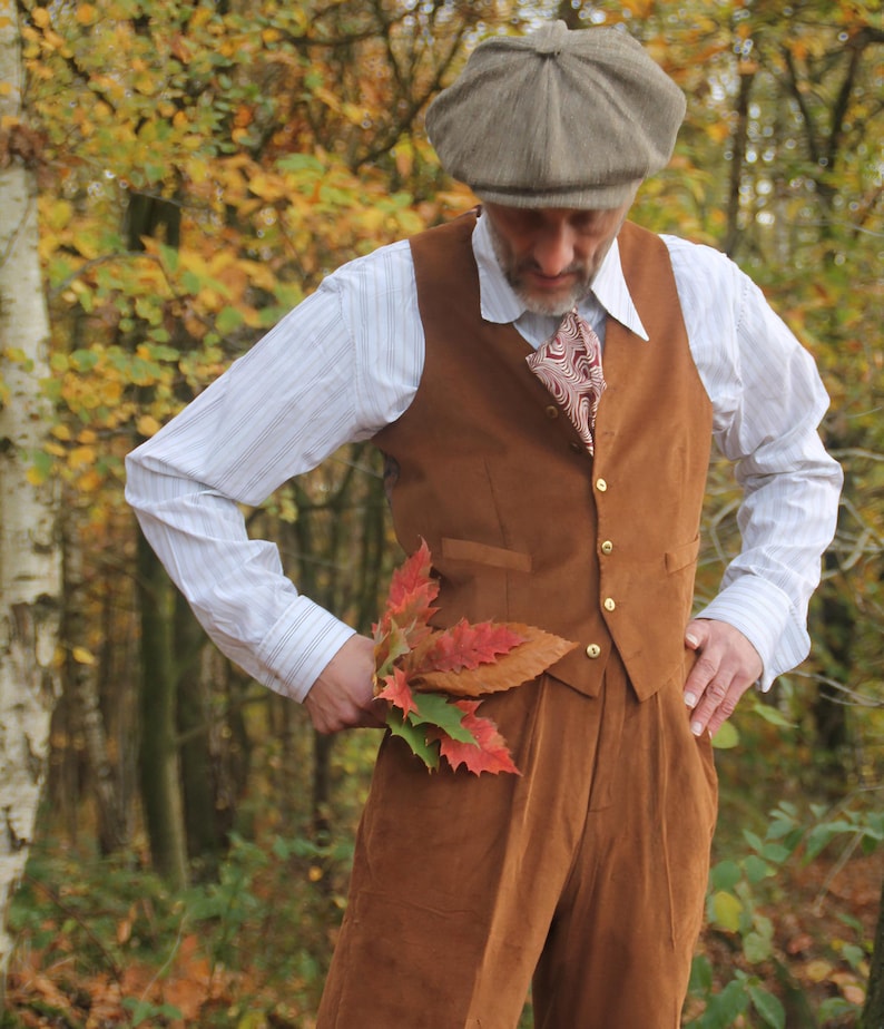 A brown suede waistcoat with gold buttons, worn with matching trousers. The outfit includes a white striped shirt, a patterned tie, and a tweed cap. The person is holding autumn leaves.