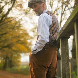 May include: A man wearing a brown tweed cap, patterned vest, and brown pants, leaning against a wooden fence. The outfit is complemented by a striped shirt. The background features a pathway and trees with autumn foliage.
