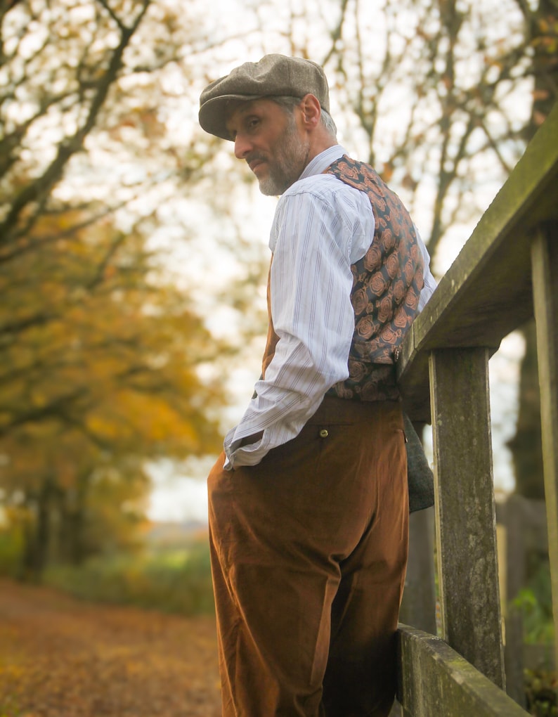 May include: A man wearing a brown tweed cap, patterned vest, and brown pants, leaning against a wooden fence. The outfit is complemented by a striped shirt. The background features a pathway and trees with autumn foliage.