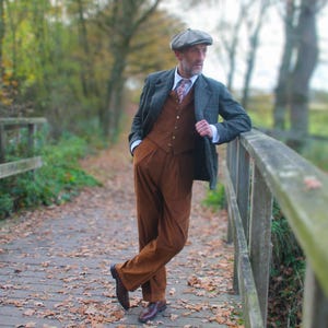 May include: A man wearing a tweed jacket, brown vest, and matching corduroy pants, with a patterned tie and a gray flat cap. He is leaning against a wooden fence on a bridge covered in fallen leaves, with a blurred background of trees.