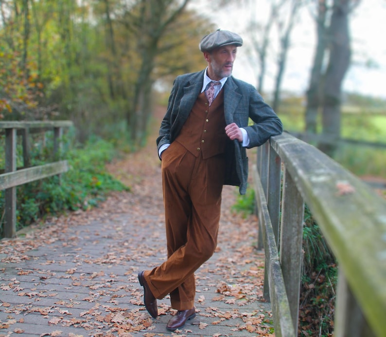 May include: A man wearing a tweed jacket, brown vest, and matching corduroy pants, with a patterned tie and a gray flat cap. He is leaning against a wooden fence on a bridge covered in fallen leaves, with a blurred background of trees.