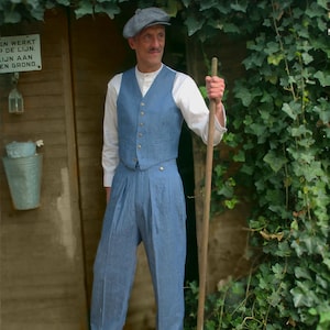 Puede incluir: Un hombre con chaleco y pantalón a rayas azules, camisa blanca de manga larga y gorra gris. Sostiene un palo de madera frente a una estructura de madera cubierta de follaje verde.