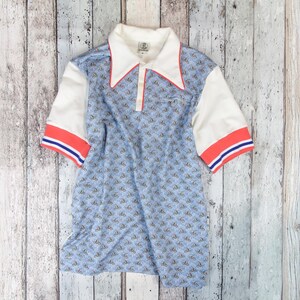 May include: A vintage polo shirt with a light blue pattern, white sleeves, and a white collar with red trim. The sleeves have red and blue stripes. The shirt is laid flat on a weathered wooden surface.