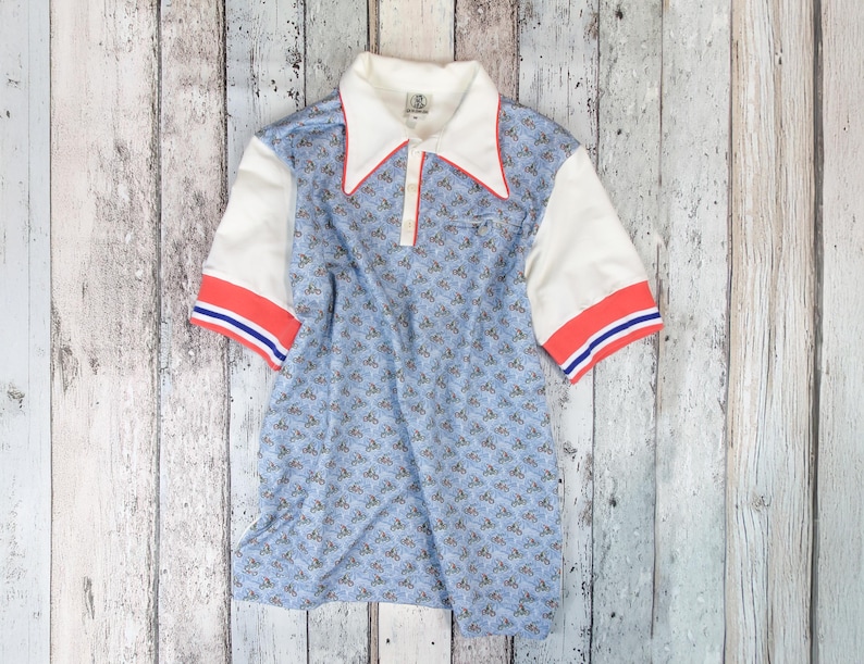 May include: A vintage polo shirt with a light blue pattern, white sleeves, and a white collar with red trim. The sleeves have red and blue stripes. The shirt is laid flat on a weathered wooden surface.