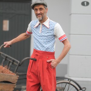 May include: A man wearing a vintage-style outfit, including a light blue polo shirt with a small pattern, red and white trim, and high-waisted red pants. He is wearing a gray cap and standing next to a bicycle.