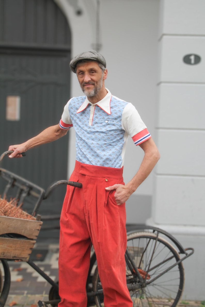 May include: A man wearing a vintage-style outfit, including a light blue polo shirt with a small pattern, red and white trim, and high-waisted red pants. He is wearing a gray cap and standing next to a bicycle.