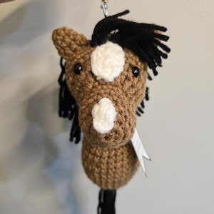 May include: A handmade crochet horse head keychain. The horse is brown with white markings on its face and a black mane and tail. It has a silver-colored clasp for attaching to keys or a bag.