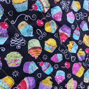May include: Black quilted fabric featuring a repeating pattern of colorful cupcakes with cherries. The cupcakes have various frosting colors, including yellow, blue, pink, and purple. White decorative elements, such as hearts and swirls, are scattered across the fabric.