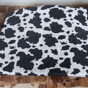May include: A rectangular, quilted fabric item with a cow print pattern. The fabric features large, irregular black spots on a white background. Horizontal stitching creates a quilted effect. The item is likely a decorative piece or a functional item.