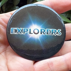 Può includere: Un bottone nero con un design a stella blu e bianca e la parola "EXPLORERS" in testo bianco.