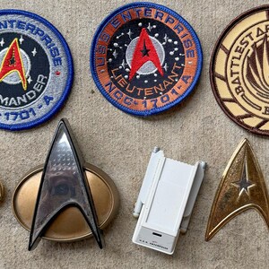 May include: A collection of science fiction memorabilia including three circular embroidered patches featuring USS Enterprise Commander, Lieutenant NCC-1701-A, and Battlestar Galactica BSG 75 designs, alongside four metallic insignia pins and a small white plastic device.