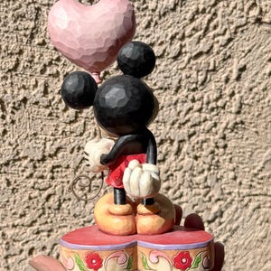 May include: A wooden Mickey Mouse figurine holding a pink heart-shaped balloon. Mickey wears red shorts, black arms, and white gloves. The figurine stands on a red base with floral detailing. The color scheme is soft and pastel, with a carved wooden texture.