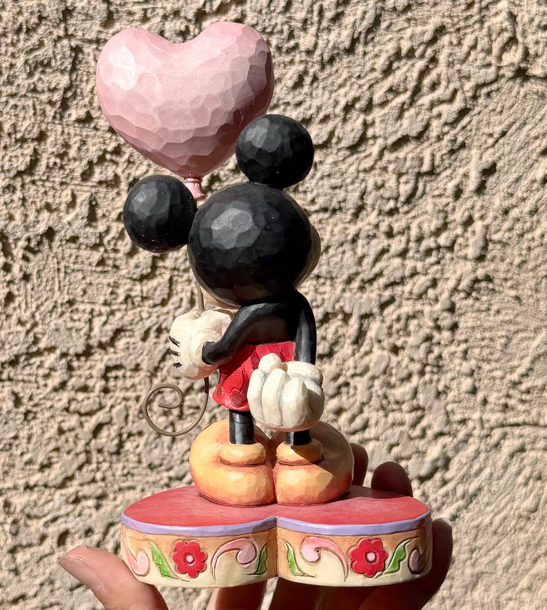 May include: A wooden Mickey Mouse figurine holding a pink heart-shaped balloon. Mickey wears red shorts, black arms, and white gloves. The figurine stands on a red base with floral detailing. The color scheme is soft and pastel, with a carved wooden texture.