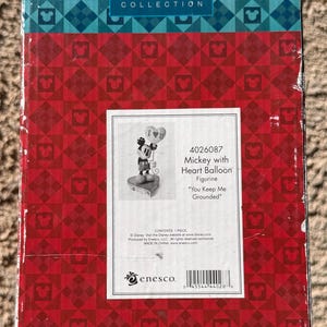 May include: A box for a Walt Disney Showcase Collection figurine. The box has a red and blue pattern with Mickey Mouse silhouettes. The label shows a Mickey Mouse figurine holding a heart balloon, with the text "You Keep Me Grounded."