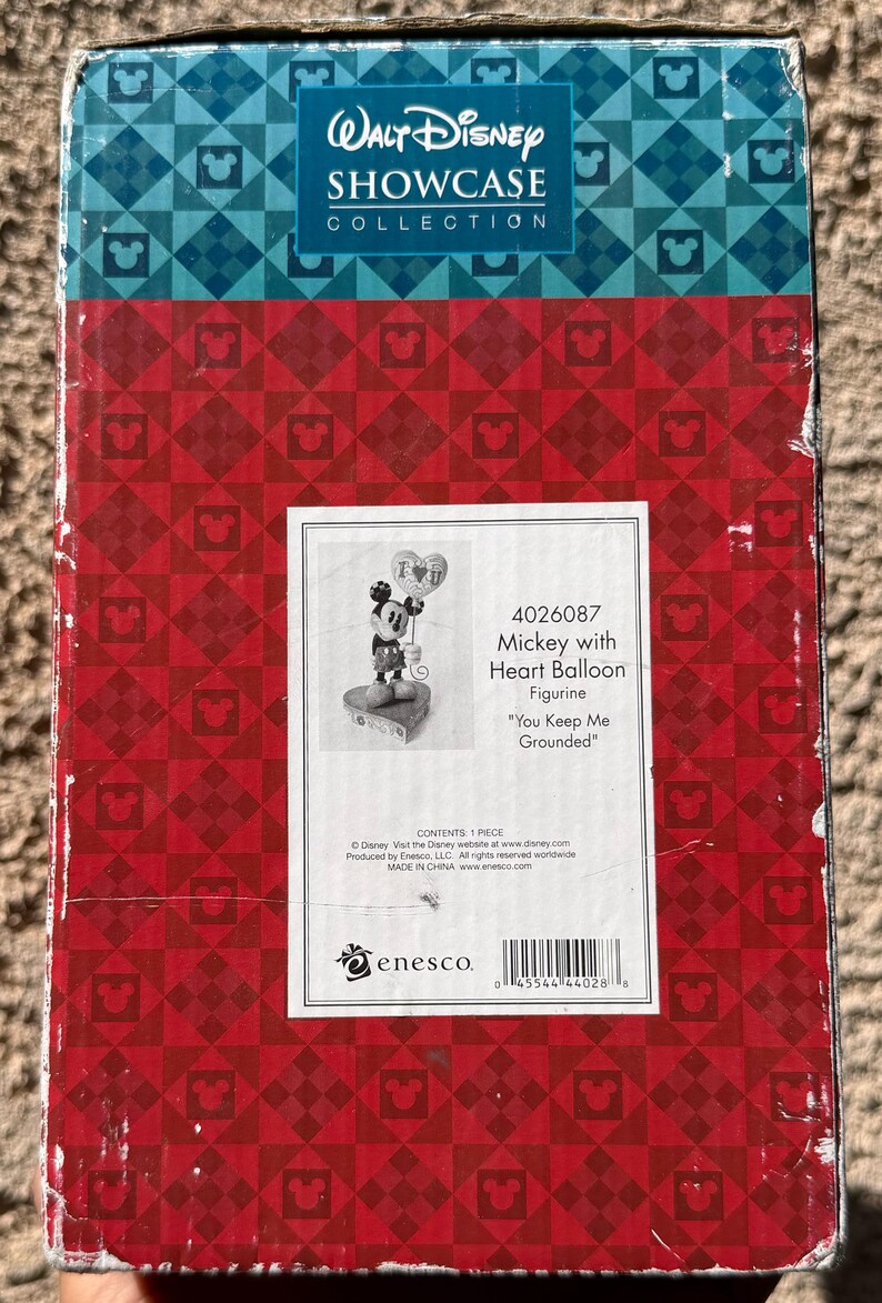 May include: A box for a Walt Disney Showcase Collection figurine. The box has a red and blue pattern with Mickey Mouse silhouettes. The label shows a Mickey Mouse figurine holding a heart balloon, with the text "You Keep Me Grounded."