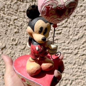May include: A Mickey Mouse figurine holding a pink heart-shaped balloon with the words "I Love You" in red. Mickey is standing on a red heart-shaped base with floral accents. The figurine is painted in bright colours.