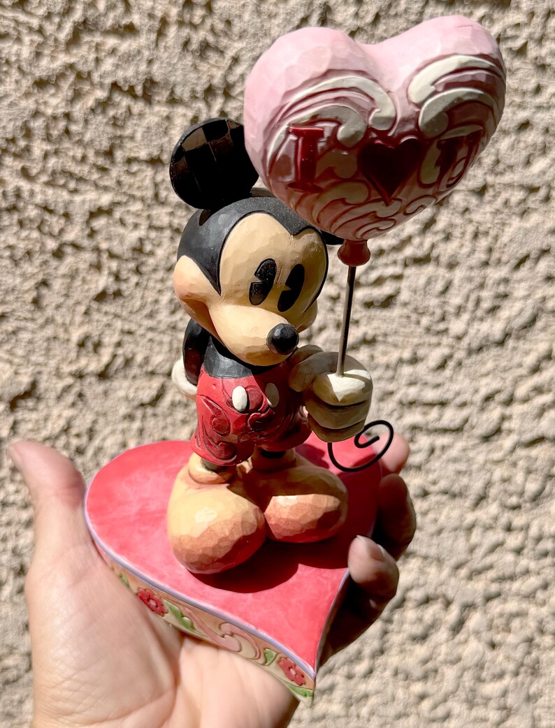 May include: A Mickey Mouse figurine holding a pink heart-shaped balloon with the words "I Love You" in red. Mickey is standing on a red heart-shaped base with floral accents. The figurine is painted in bright colours.