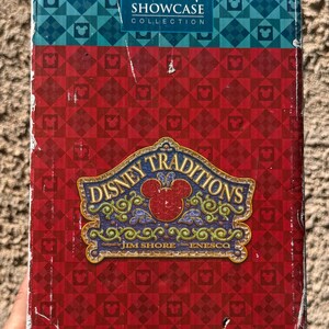 May include: A box with the "Walt Disney Showcase Collection" logo in white on a blue and teal patterned section. The box has a red patterned background with the "Disney Traditions" logo in the centre, featuring a Mickey Mouse head.