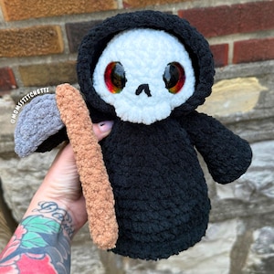May include: A plush crochet grim reaper doll featuring a white skull face with large red and orange eyes, wearing a black hooded robe. The figure holds a scythe with a grey blade and a tan handle. The text @MOMSSTITCHETTI is visible on the left.