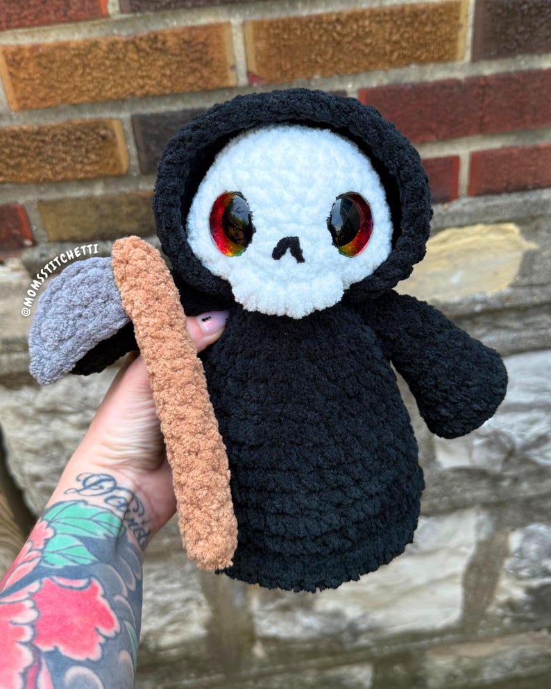 May include: A plush crochet grim reaper doll featuring a white skull face with large red and orange eyes, wearing a black hooded robe. The figure holds a scythe with a grey blade and a tan handle. The text @MOMSSTITCHETTI is visible on the left.