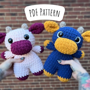 May include: Two crocheted stuffed cow toys. One is pink with a purple nose and ears, the other is blue with a yellow nose and ears. Both have large black eyes. The toys are held in the hands of a person with tattoos.
